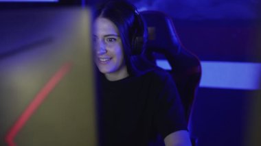 Güzel bir genç kadın kulaklıkla oturur ve bir bilgisayar kulübünde online oyun oynar. Kadın hevesle partnerleriyle mikrofon başında oynuyor ve konuşuyor. Dikey görünüm.