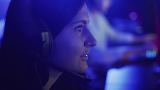 Hermosa mujer juega un juego en línea en un moderno club de informática. Mujer joven se sienta en los auriculares delante del monitor, mira con atención y se ríe. Emociones de alegría y sorpresa en un juego de ordenador — Vídeos de Stock