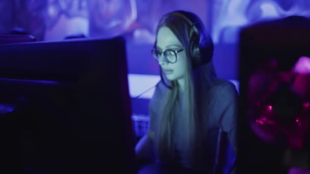 Mujer joven concentrada con gafas y auriculares se sienta frente al monitor y juega en el videojuego en el club de computadoras — Vídeos de Stock