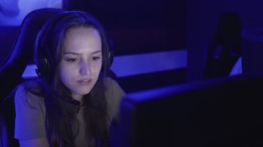 Genç bir kadın modern bir bilgisayar kulübünde kulaklıkla oturup online oyun oynuyor. Kadın gülümsüyor ve internet oyununa şaşırıyor. Dikey görünüm.