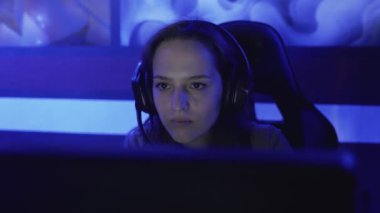 Genç bir kadın modern bir bilgisayar kulübünde kulaklıkla oturup online oyun oynuyor. Kadın gülümsüyor ve internet oyununa şaşırıyor. Dikey görünüm.