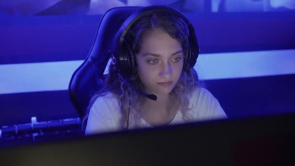 Mujer joven se sienta en los auriculares en un club de informática moderna, juega un juego en línea. Mujer sonríe y se sorprende con el juego de Internet. Vista de retrato. — Vídeos de Stock