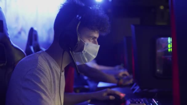 Vue latérale du jeune homme dans le masque facial joue dans le jeu vidéo en ligne dans le club informatique moderne. — Video
