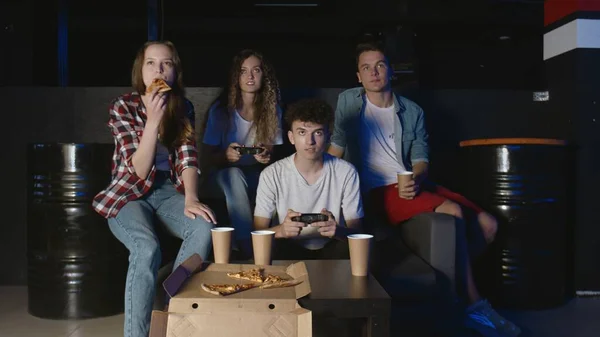 Jovens homens e mulheres alegres estão se divertindo com o videogame na festa em casa enquanto os amigos riem assistindo, conversando e comendo pizza — Fotografia de Stock