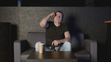 Genç adam kameranın önünde konsol oynuyor, rakibini internette yeniyor. Kanepede beyaz bir adam.