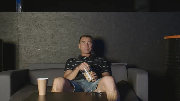 Adult man sits on couch, eats popcorn and watches TV