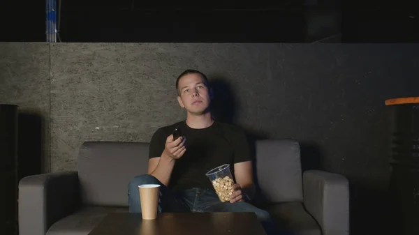 Красивый человек ест попкорн и смотрит телевизор дома с помощью пульта дистанционного управления расслабляясь на диване — стоковое фото