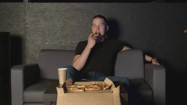 Çekici bir adam pizza yiyor ve evde televizyon seyrediyor. Kanepede uzaktan kumandayla dinleniyor. — Stok fotoğraf