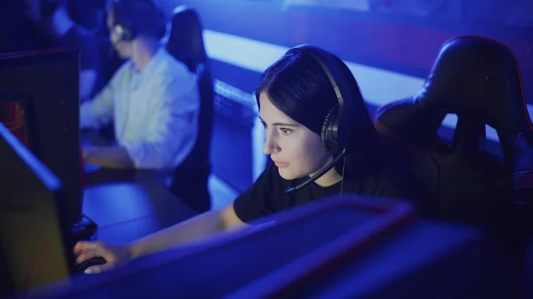 Hermosa mujer juega un juego en línea en un moderno club de informática. Mujer joven se sienta en los auriculares delante del monitor, mira con atención y se ríe. —  Fotos de Stock