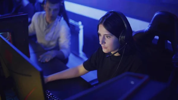 Mujer joven se sienta en los auriculares en un club de informática moderna, juega un juego en línea. Mujer sonríe y se sorprende con el juego de Internet. Vista de retrato. — Foto de Stock