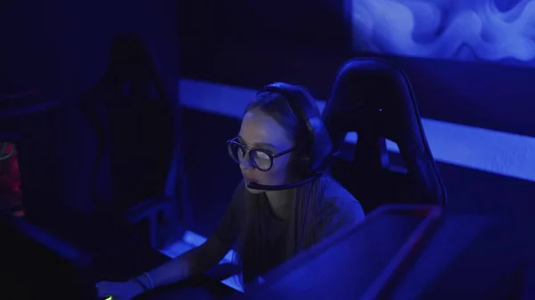 Mujer joven concentrada con gafas y auriculares se sienta frente al monitor y juega en el videojuego en el club de computadoras —  Fotos de Stock