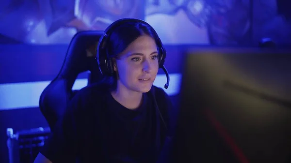 Jeune femme s'assoit dans un casque dans un club informatique moderne, joue à un jeu en ligne. Femme sourit et est surprise par le jeu Internet. Vue portrait. — Photo