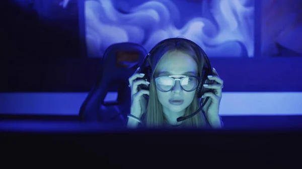 Concentrated young woman with glasses and headphones sits in front of monitor and plays in video game in computer club Royalty Free Stock Photos