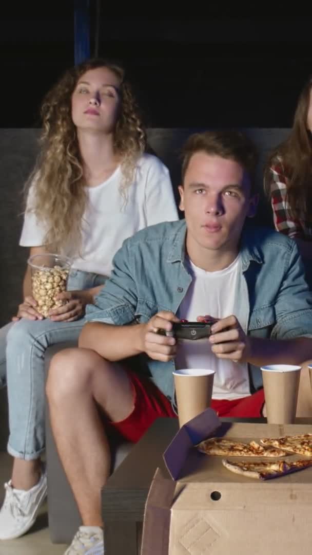 Młodzi przyjaciele dwóch facetów i dwie dziewczyny grają na konsoli. Siedzą na kanapie i trzymają joysticki. VIDEO WERTYCZNE — Wideo stockowe