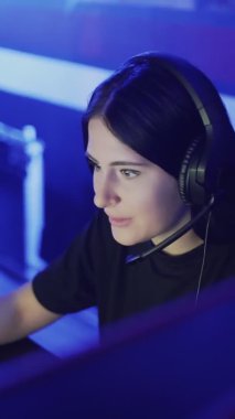 Güzel bir kadın modern bir bilgisayar kulübünde online oyun oynuyor. Genç kadın kulaklıkla monitörün önünde oturuyor, dikkatli bakıyor ve gülüyor. VERTICAL VIDEO