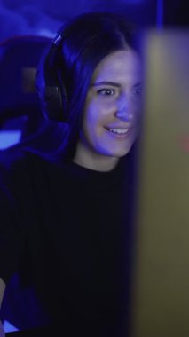 Güzel bir genç kadın kulaklıkla oturur ve bir bilgisayar kulübünde online oyun oynar. Kadın hevesle partnerleriyle mikrofon başında oynuyor ve konuşuyor. Portre görüntüsü. VERTICAL VIDEO