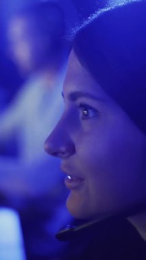 Güzel bir kadın modern bir bilgisayar kulübünde online oyun oynuyor. Genç kadın kulaklıkla monitörün önünde oturuyor, dikkatli bakıyor ve gülüyor. VERTICAL VIDEO