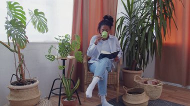 Güzel Afro-Amerikalı kadın etrafı çiçeklerle çevrili, hasır sandalyede oturur, çay içer ve kitap okur.