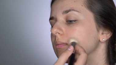 Stüdyoda oturan güzel ve muhteşem kadın için profesyonel makyaj süreci. Makyaj sanatçısı makyaj fırçası kullanarak cilt bakım kremi sürüyor