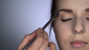 Stüdyoda oturan güzel ve göz kamaştırıcı kadın için Profesyonel Makyaj Süreci 'ni kapat. Makyaj sanatçısı göz kapağındaki fırçayla allık sürüyor