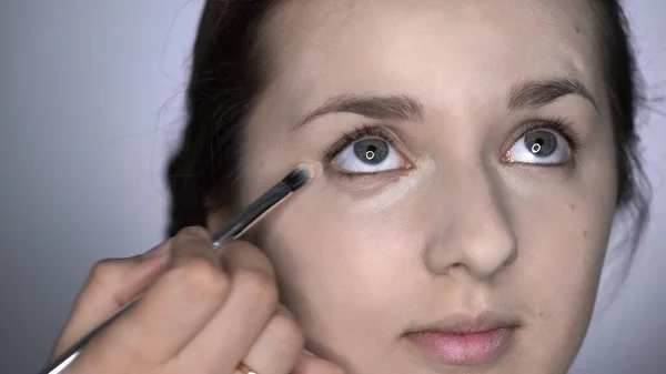 Processo de Maquiagem Profissional para mulher bonita e linda sentada no estúdio. Maquiagem Artista aplica corretor na área dos olhos — Fotografia de Stock