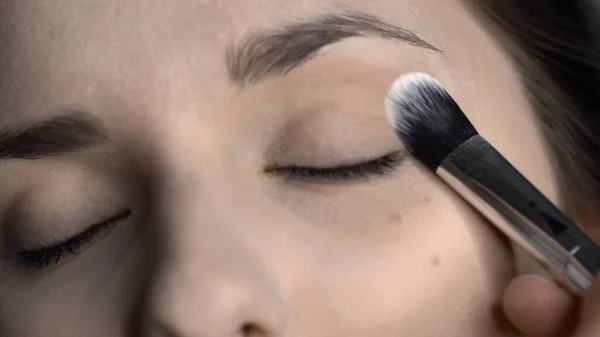 Proceso de maquillaje profesional para una mujer hermosa y hermosa sentada en el estudio. Maquillaje Artista aplica polvo con un cepillo grande en el área de los ojos — Foto de Stock