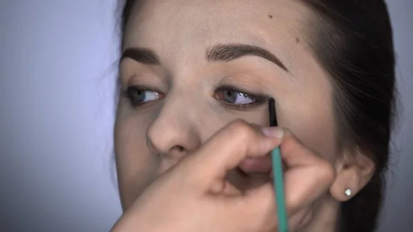 Proces profesjonalnego makijażu dla pięknej i wspaniałej kobiety siedzącej w studio. Make up Artysta stosując eyeliner na górnej powiece brązowookiej dziewczyny — Zdjęcie stockowe