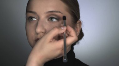 Güzellik stüdyosunda genç bir kadın için profesyonel makyaj yapıyor. Makyaj sanatçısı göz kapağına gölge uygulamak için fırça kullanır