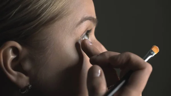 Vista lateral del maquillador haciendo maquillaje profesional para mujer joven en el estudio de belleza. Maquillaje Artista aplica corrector en el área de ojos — Foto de Stock