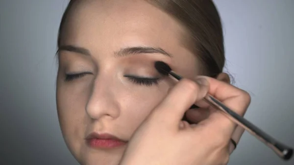 Makijażysta robi profesjonalny makijaż dla młodej kobiety w studiu kosmetycznym. Make up Artist używa pędzla do nakładania cienia na powiekę — Zdjęcie stockowe