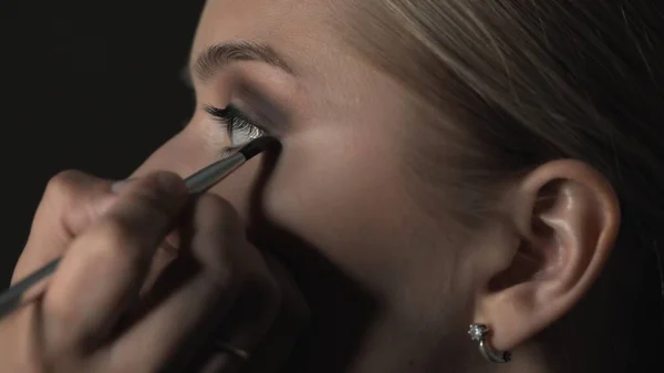 Vista lateral del maquillador haciendo maquillaje profesional para mujer joven en el estudio de belleza. Maquillaje Artista utiliza cepillo para aplicar sombra en el párpado — Foto de Stock