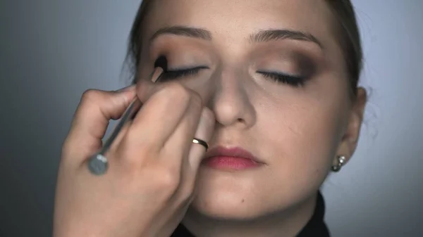 Vista frontal del maquillador haciendo maquillaje profesional para mujer joven en el estudio de belleza. Maquillaje Artista utiliza cepillo para aplicar sombra en el párpado — Foto de Stock