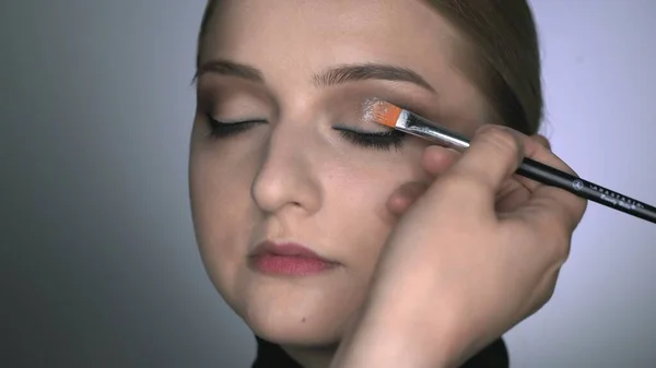 Maquillaje artista haciendo maquillaje profesional para mujer joven en el estudio de belleza. Maquillaje Artista utiliza cepillo para aplicar brillo en los ojos — Foto de Stock