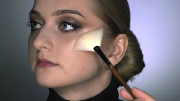 Макияж художник делает профессиональный макияж для молодой женщины в студии красоты. Макияж Artist использует кисть для нанесения глайтера или хайлайтера — стоковое фото
