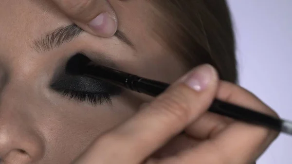 Makijażysta robi profesjonalny makijaż dla młodej kobiety w studiu kosmetycznym. Make up Artist sprawia, że makijaż smokey oczy czarny odcień ze specjalnym pędzlem piękny model — Zdjęcie stockowe