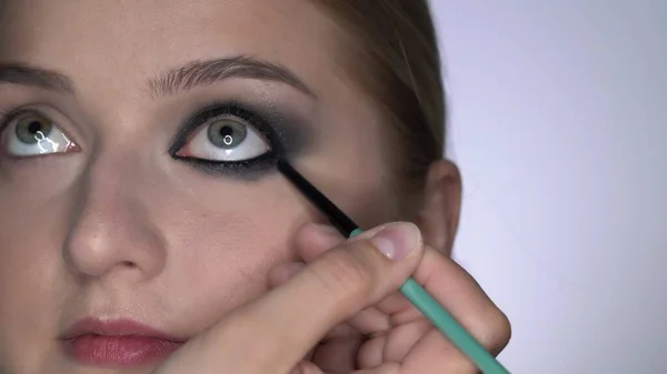 Makijażysta robi profesjonalny makijaż dla młodej kobiety w studiu kosmetycznym. Make up Artist sprawia, że makijaż smokey oczy czarny odcień ze specjalnym pędzlem piękny model — Zdjęcie stockowe