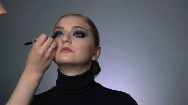 Vista lateral do artista de maquiagem fazendo maquiagem profissional para a jovem no estúdio de beleza. Maquiagem Artista faz a maquiagem olhos esfumaçados de uma sombra preta com escova especial de um belo modelo — Fotografia de Stock