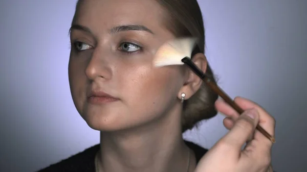 Maquillaje artista haciendo maquillaje profesional para mujer joven en el estudio de belleza. Maquillaje Artista utiliza cepillo para aplicar brillo o iluminador —  Fotos de Stock