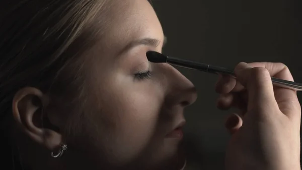 Side view van make-up kunstenaar het maken van professionele make-up voor jonge vrouw in beauty studio. Make-up Kunstenaar gebruikt penseel om schaduw aan te brengen op ooglid Rechtenvrije Stockafbeeldingen