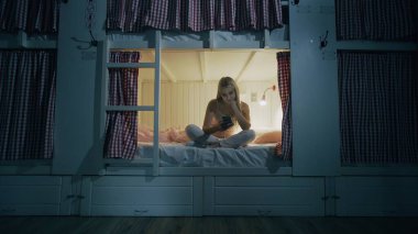 Yurt odasındaki yatakta oturan ve gece akıllı telefon kullanan mutlu kadın.