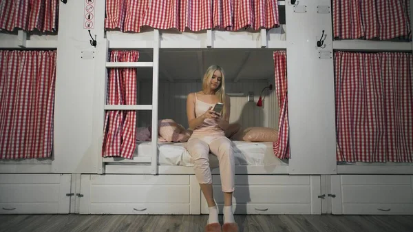 Junge Frau sitzt im Hostel-Zimmer auf dem Bett und benutzt Smartphone — Stockfoto
