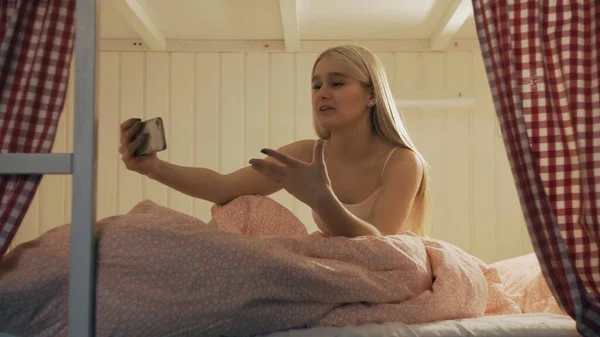 Close up of young attractive woman sitting on the bed in the hostel room and using smartphone having video chat — Stock Photo, Image