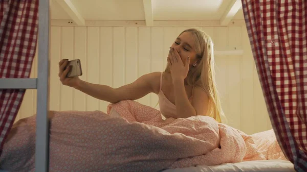 Gros plan de la jeune femme attrayante assise sur le lit dans la salle de l'auberge et en utilisant smartphone ayant chat vidéo — Photo