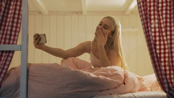 Close up of young attractive woman sitting on the bed in the hostel room and using smartphone having video chat Stock Image