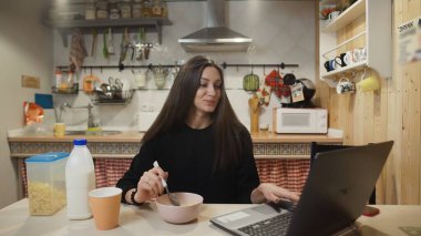 Dizüstü bilgisayarda çalışan ve mısır gevreği yiyen genç bir kadın..