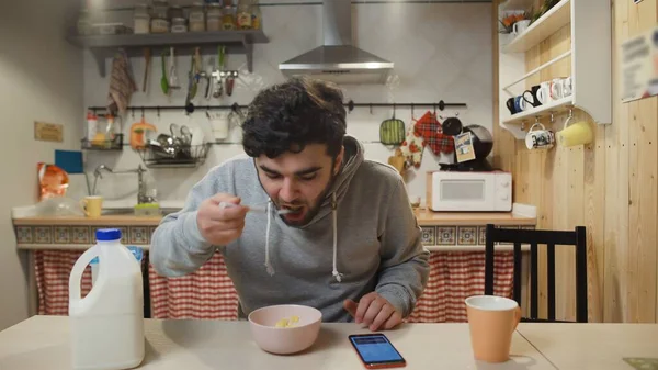 Молодой человек завтракает на кухне. Человек ест зерновые хлопья и использует смартфон — стоковое фото