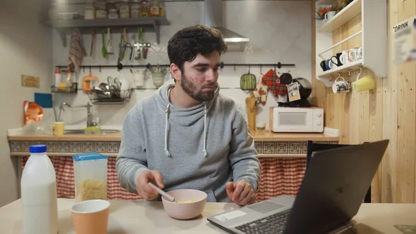 Young man working laptop computer and eats Corn Flakes Cereal at home kitchen. — Stock Photo, Image