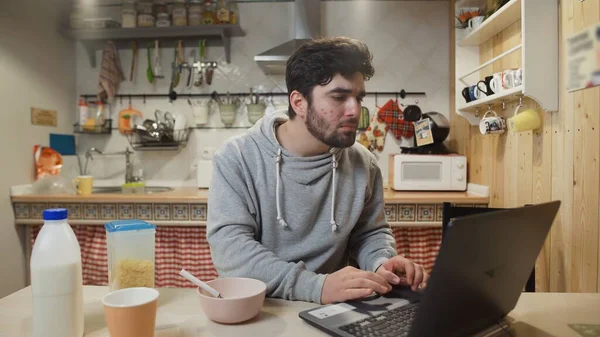 Genç adam dizüstü bilgisayarda çalışıyor ve mısır gevreği yiyor..