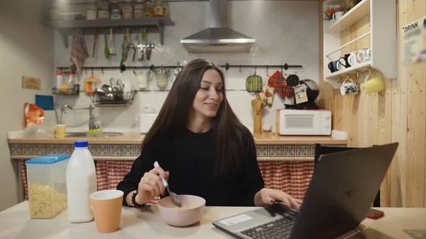Junge Frau arbeitet am Laptop und isst Cornflakes-Müsli in der heimischen Küche. — Stockfoto