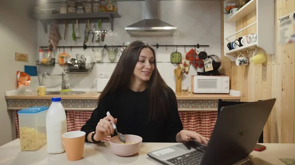 Young woman working laptop computer and eats Corn Flakes Cereal at home kitchen. — Stock Photo, Image